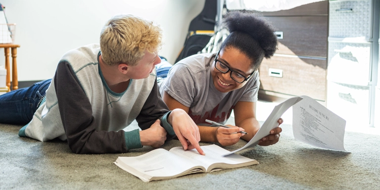 A male and female student lay on the floor studying with books and papers in front of them.