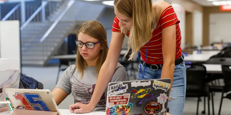 A female student stands beside a seated female student providing math tutoring in a library 