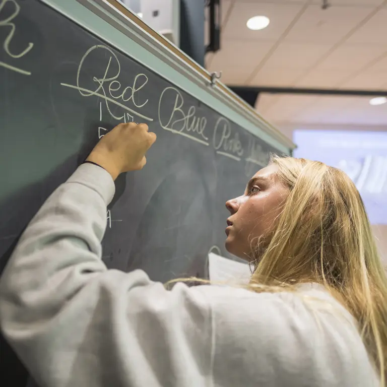 Left-handed female student writing on a green chalkboard