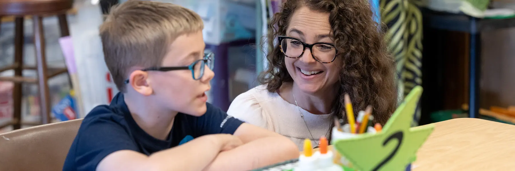 A female teacher crouches next to a young male student working on an art project at a classroom table