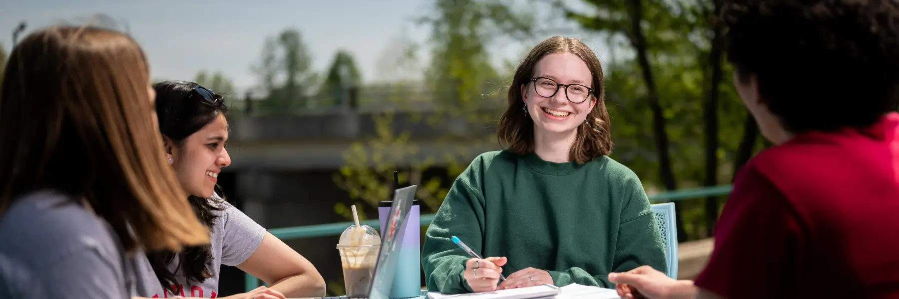 A female student seated at an outdoor table smiles at her friends as they study