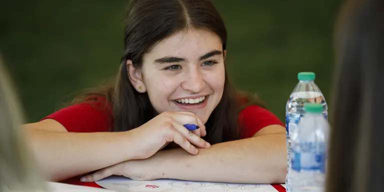 A female student smiles and rests her chin on crossed arms over an outdoor table.