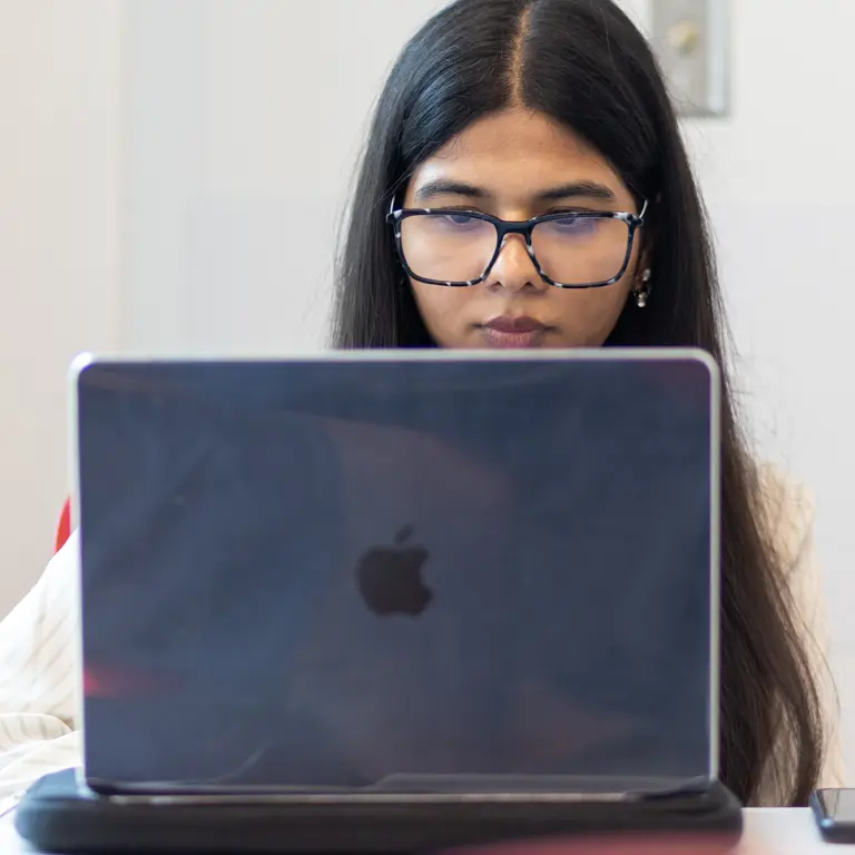 A female student with long black hair and glasses works on a laptop