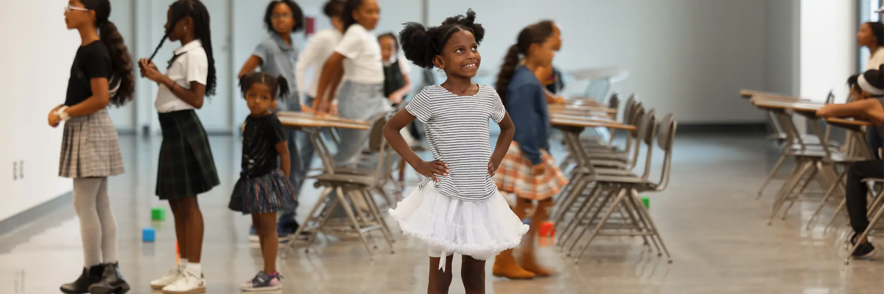 A small female child wearing a tutu stands with her hands on her hips in a busy classroom