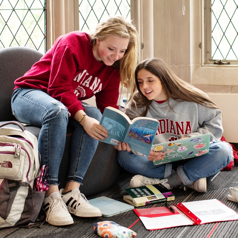 A female student in a chair leans to a friend seated on the floor to show a page in a book. 