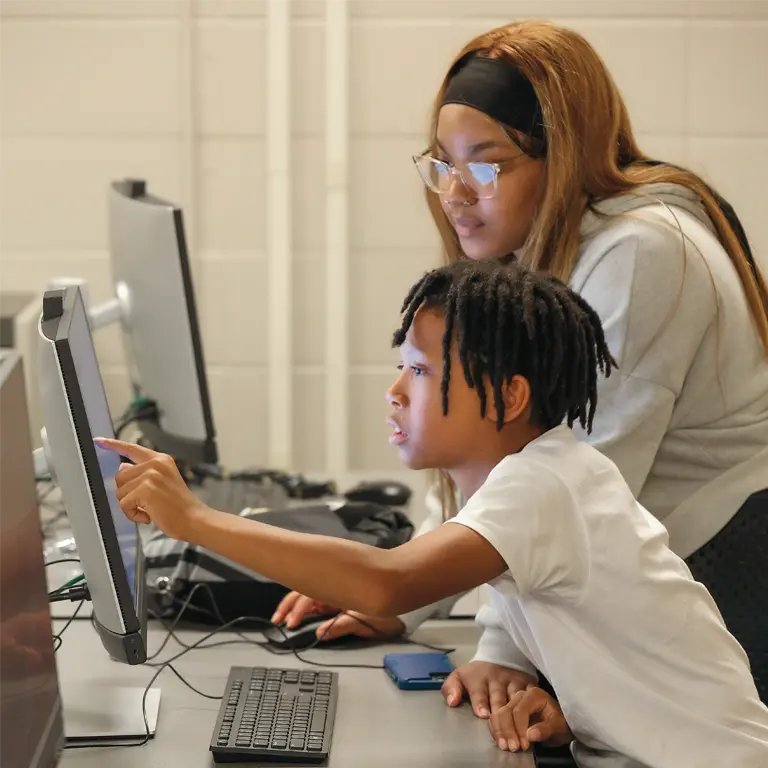 A Kids College instructor works with a young male participant at a desktop computer