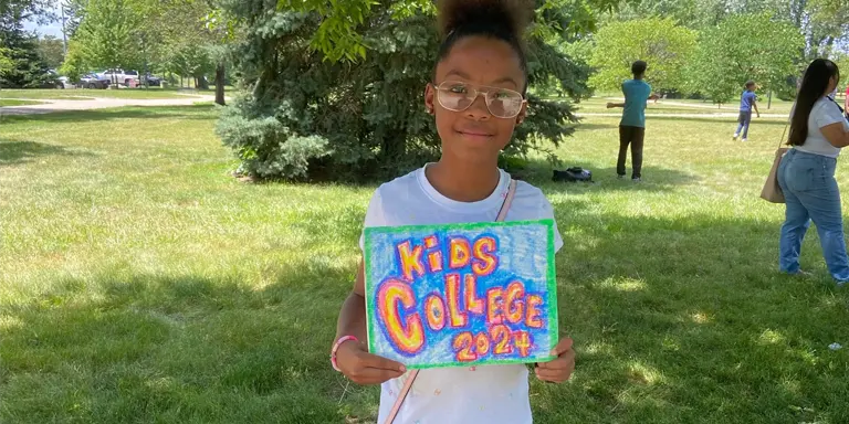 A young female participant holds a handmade sign that reads Kids College