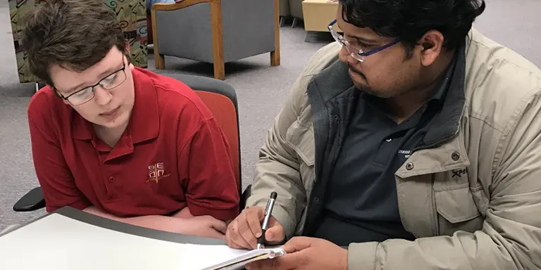 A male student is seated beside another male student in a library providing math tutoring 