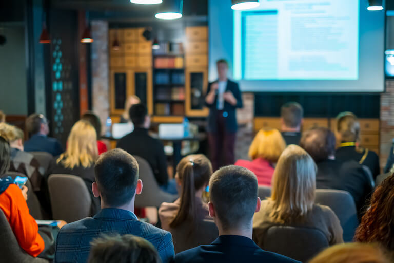 Teachers sitting in a large classroom paying attention to a presenter with a powerpoint presentation in the front of the room.